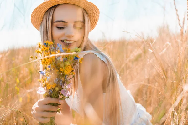 Selective focus of sensual woman in straw hat holding wildflowers in grassy field — Stock Photo