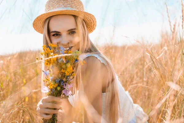 Selektiver Fokus einer jungen Frau mit Strohhut, die Wildblumen in der Hand hält und auf der Wiese in die Kamera blickt — Stockfoto