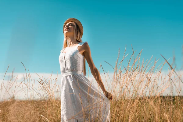 Selective focus of young woman in straw hat touching white dress while standing with closed eyes in grassy meadow — Stock Photo