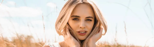 Conceito panorâmico de mulher loira pensativa tocando o cabelo enquanto olha para a câmera contra o céu azul — Fotografia de Stock