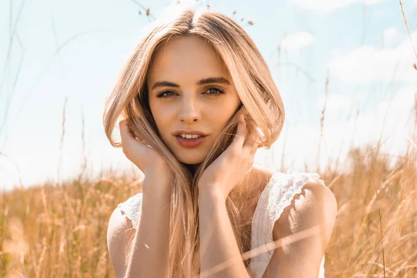 Messa a fuoco selettiva della donna bionda sognante che tocca i capelli e guarda la fotocamera contro il cielo blu nel prato — Foto stock