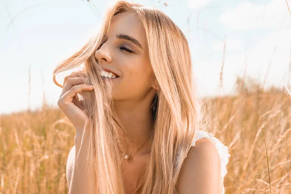 Femme blonde sensuelle touchant les cheveux et regardant loin près de l'herbe dans le champ — Photo de stock