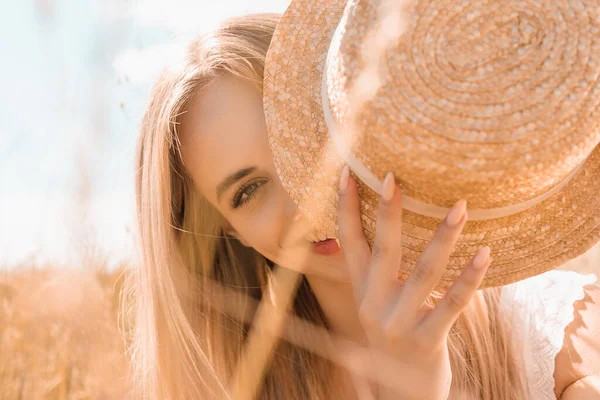 Selektiver Fokus der blonden Frau, die ihr Gesicht mit Strohhut verdeckt, während sie in die Kamera blickt — Stockfoto