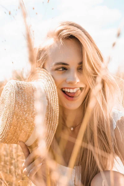 Вибірковий фокус блондинки, що стирчить язиком, тримаючи солом'яний капелюх і дивлячись в поле — стокове фото