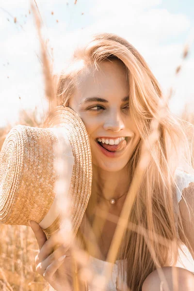 Selective focus of blonde woman sticking out tongue while holding straw hat and looking away in field — Stock Photo