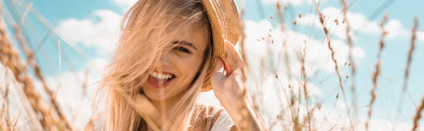 Selektiver Fokus der aufgeregten blonden Frau, die Strohhut berührt und vor bewölktem Himmel in die Kamera blickt, horizontales Bild — Stockfoto