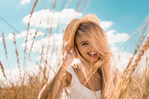 Foyer sélectif de femme blonde excitée touchant chapeau de paille et regardant caméra contre ciel nuageux bleu — Photo de stock