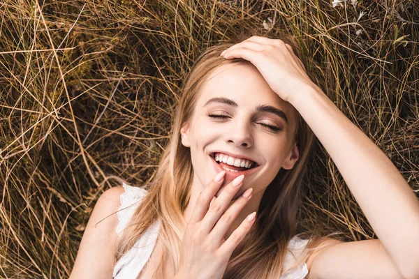 Vista superior de la mujer rubia emocionada tocando los labios y la frente mientras está acostado en la hierba con los ojos cerrados - foto de stock