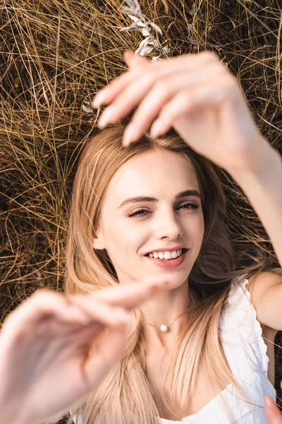 Draufsicht der aufgeregten blonden Frau, die mit erhobenen Händen im Gras liegt und in die Kamera blickt — Stockfoto