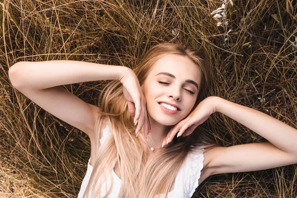 Vue de dessus de la femme blonde sensuelle touchant le visage tout en étant couché sur l'herbe verte avec les yeux fermés — Photo de stock