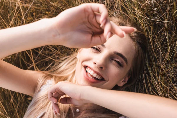 Vue de dessus de femme blonde excitée allongée sur l'herbe avec les mains levées et regardant la caméra, mise au point sélective — Photo de stock