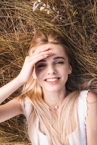 Vista superior da mulher loira animado tocando na testa e olhando para a câmera enquanto deitado na grama verde — Fotografia de Stock