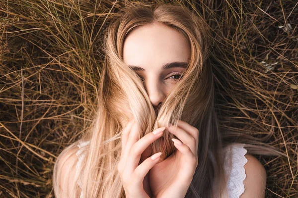 Vue de dessus de la jeune femme blonde obscurcissant le visage avec des cheveux tout en étant allongé sur l'herbe et en regardant la caméra — Photo de stock