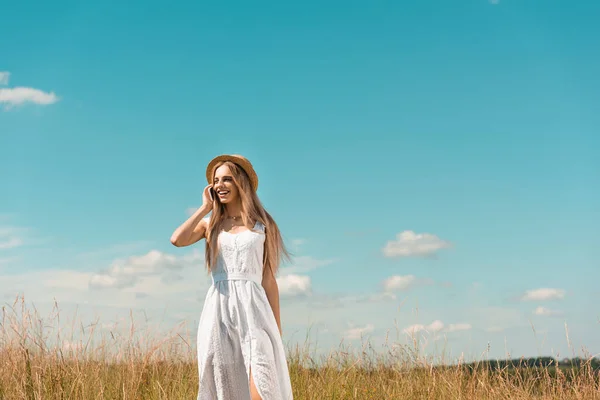 Stylish blonde woman in white dress and straw hat talking on smartphone in meadow against blue sky — Stock Photo