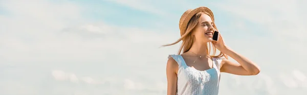 Plano panorámico de mujer rubia en sombrero de paja hablando en smartphone con los ojos cerrados contra el cielo azul - foto de stock