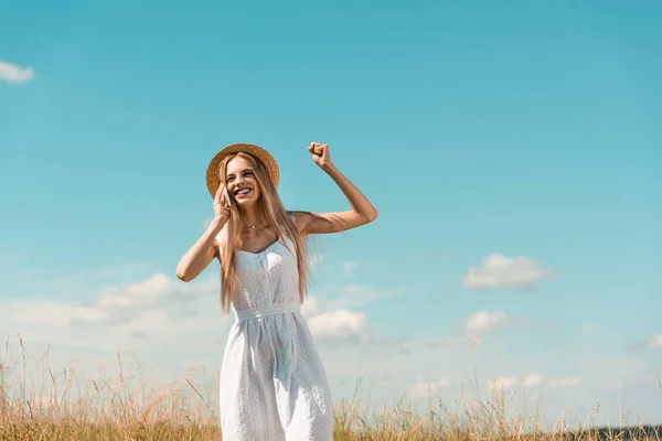 Aufgeregte blonde Frau in weißem Kleid und Strohhut zeigt Siegergeste, während sie mit dem Smartphone vor blauem Himmel spricht — Stockfoto
