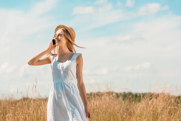 Junge Frau in weißem Kleid und Strohhut spricht mit Smartphone, während sie mit geschlossenen Augen auf dem Feld steht — Stockfoto