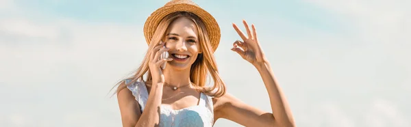 Horizontales Bild einer blonden Frau mit Strohhut, die bei Gesprächen auf dem Smartphone vor blauem Himmel eine gute Geste zeigt — Stockfoto