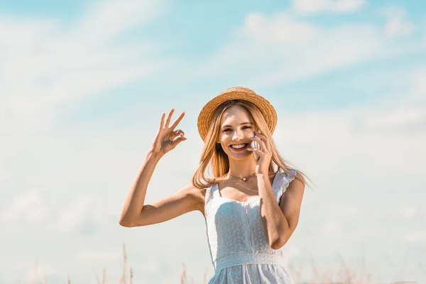 Jovem, mulher animada em vestido branco e chapéu de palha mostrando gesto ok enquanto fala no smartphone contra o céu azul — Fotografia de Stock