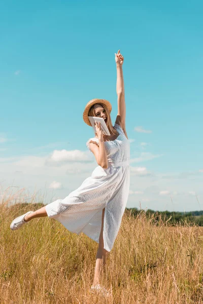 Збуджена блондинка в білій сукні говорить на цифровому планшеті, стоячи на одній нозі з піднятою рукою проти блакитного неба — стокове фото