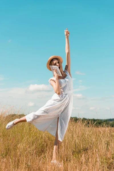 Mujer rubia excitada en vestido blanco hablando en tableta digital mientras está de pie en una pierna con la mano levantada contra el cielo azul - foto de stock