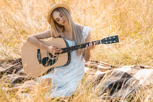 Donna sognante in abito bianco e cappello di paglia suonare la chitarra acustica mentre seduto su una coperta in campo — Foto stock