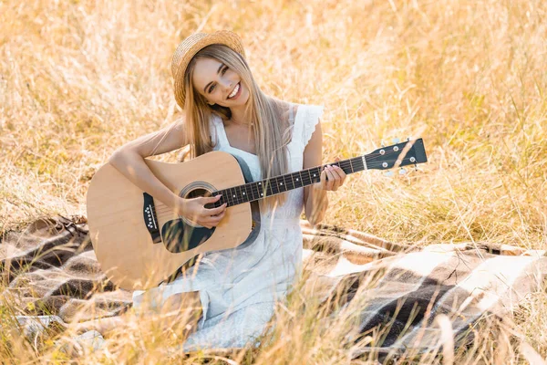 Selektiver Fokus einer jungen Frau in weißem Kleid und Strohhut, die in die Kamera schaut, während sie auf einer Decke im Feld Akustikgitarre spielt — Stockfoto