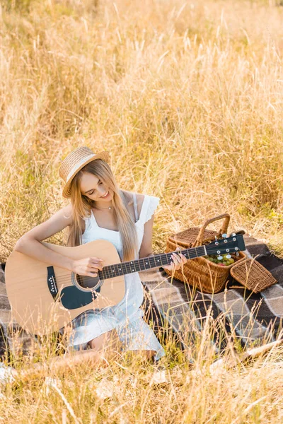 Vista ad alto angolo della donna bionda in abito bianco e cappello di paglia che suona la chitarra acustica sulla coperta in campo, messa a fuoco selettiva — Foto stock