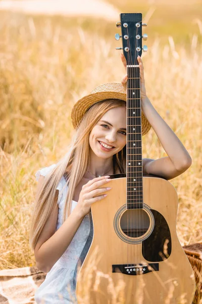 Foco seletivo de jovem loira olhando para a câmera enquanto sentado com guitarra acústica em campo gramado — Fotografia de Stock