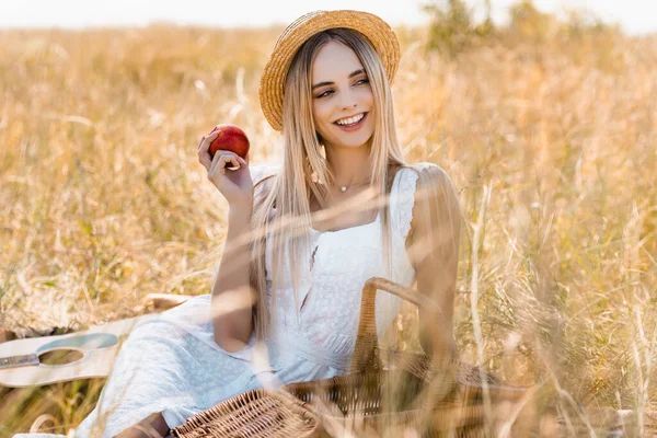 Selective focus of woman in white dress and straw hat holding ripe apple while sitting in field — Stock Photo