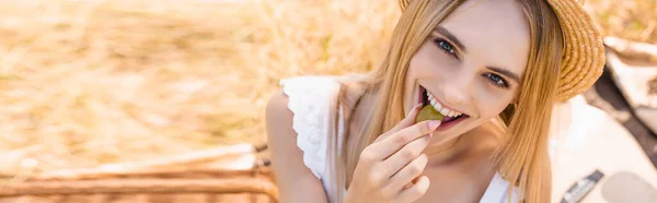 Horizontal image of young woman in summer outfit eating ripe grape and looking at camera in meadow — Stock Photo