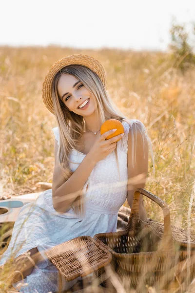 Enfoque selectivo de la mujer con estilo en vestido blanco y sombrero de paja sosteniendo naranja mientras está sentado en el campo cerca de la cesta de mimbre - foto de stock