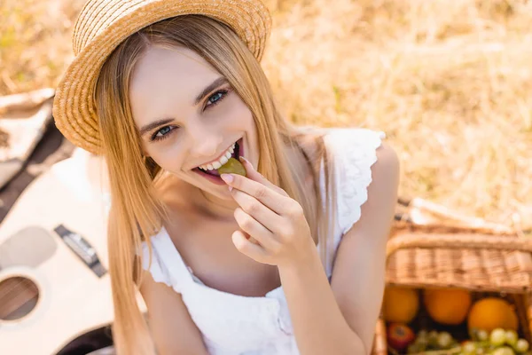 Vue grand angle de femme blonde en chapeau de paille mangeant du raisin mûr et regardant la caméra sur pique-nique dans le champ — Photo de stock
