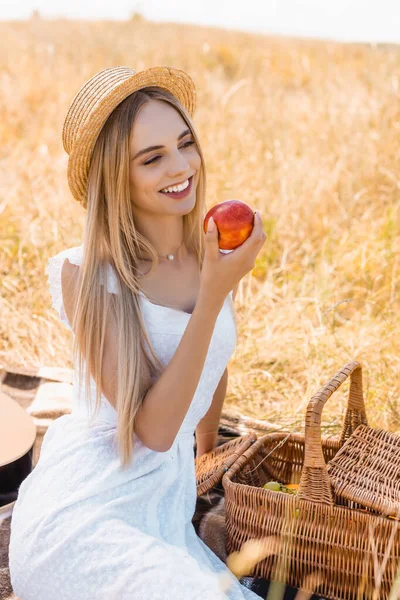 Enfoque selectivo de la mujer rubia en sombrero de paja y vestido blanco sentado en la manta cerca de la canasta de mimbre y la celebración de la manzana madura - foto de stock