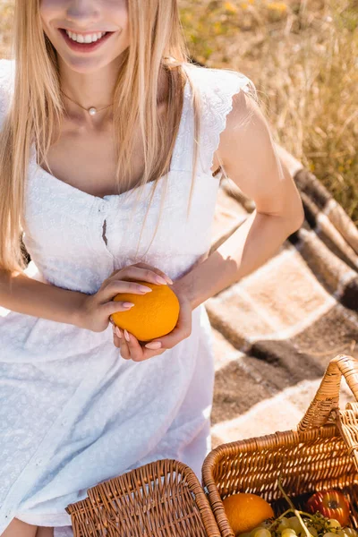 Vue recadrée de la femme en robe blanche tenant orange tout en étant assis sur la couverture près du panier en osier — Photo de stock