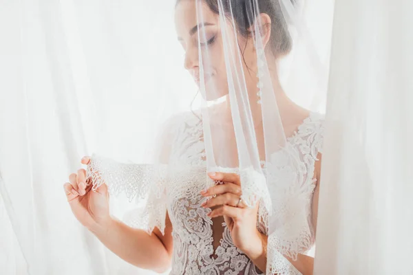 Selective focus of young bride holding lace veil near white curtains — Stock Photo