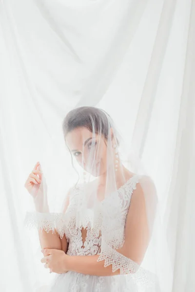 Невеста в свадебном платье и вуаль, смотрящая в камеру возле белой ткани — стоковое фото