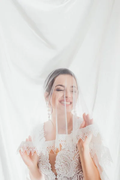 Brünette Braut im Brautkleid berührt Spitzenschleier in der Nähe von weißem Stoff — Stockfoto