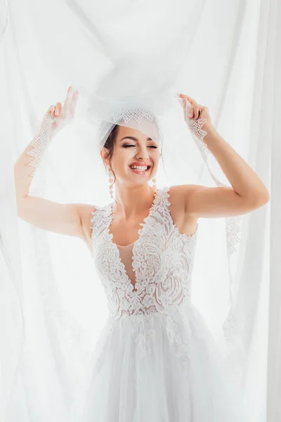 Braut im Brautkleid aus Spitze hält Schleier in der Nähe von weißem Tuch — Stockfoto