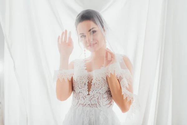 Junge Braut in Brautkleid und Schleier blickt in die Kamera neben weißen Vorhängen — Stockfoto