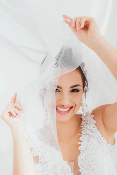 Selective focus of bride in wedding dress holding veil and looking at camera near white cloth — Stock Photo