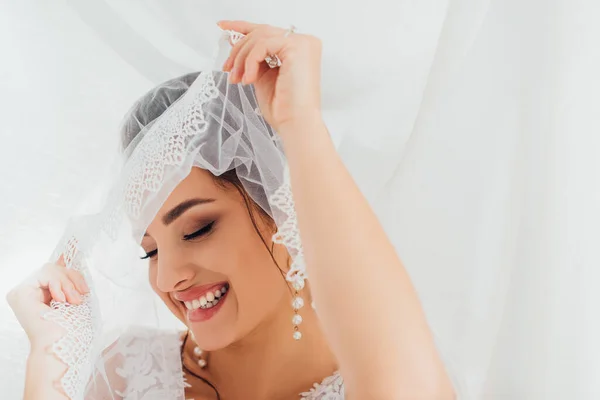 Выборочный фокус невесты в жемчужных серьгах держа кружевную вуаль возле белой ткани — стоковое фото