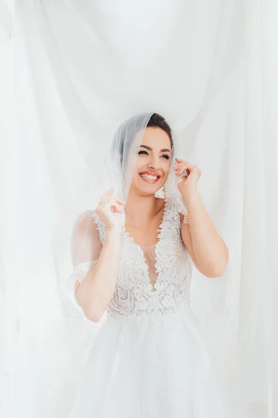 Morena novia en vestido de novia y velo mirando hacia otro lado cerca de tela blanca - foto de stock