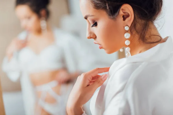 Enfoque selectivo de la novia en pendientes de perlas tocando la túnica de seda en casa - foto de stock
