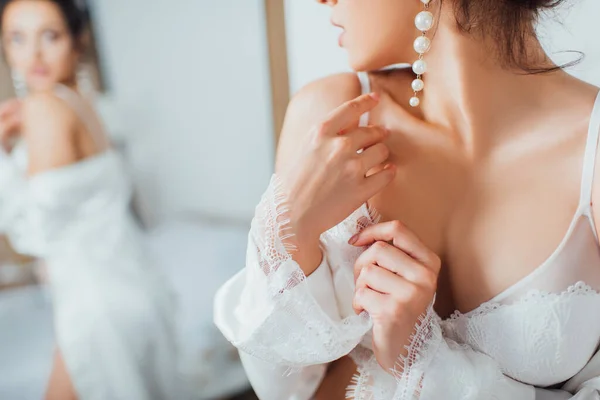 Vista ritagliata della sposa in reggiseno di pizzo e orecchino di perle toccare accappatoio di seta a casa — Foto stock