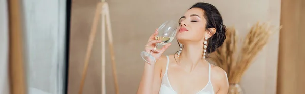 Panoramic shot of bride in bra and pearl earrings holding glass of wine at home — Stock Photo