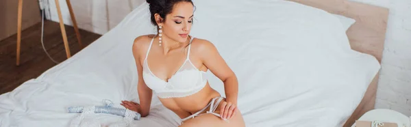 Panoramic concept of sexy bride touching garter belt while sitting on bed — Stock Photo