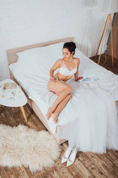 Sexy bride in lingerie touching garter belt near wedding dress on bed at home — Stock Photo