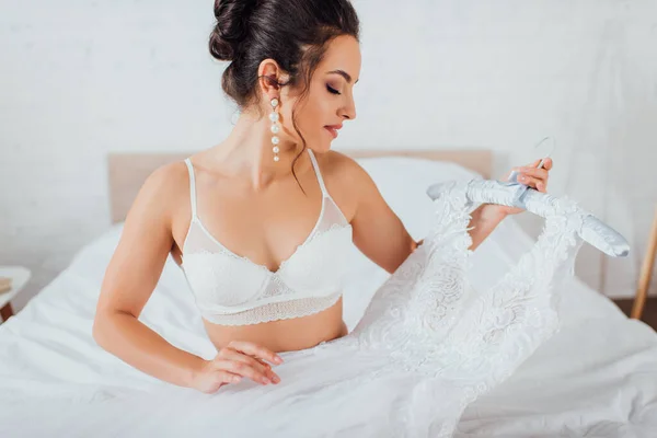 Brünette Braut in Spitzen-BH hält Brautkleid auf Kleiderbügel im Schlafzimmer — Stockfoto