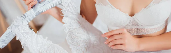 Panoramic shot of bride in bra holding wedding dress on hanger at home — Stock Photo